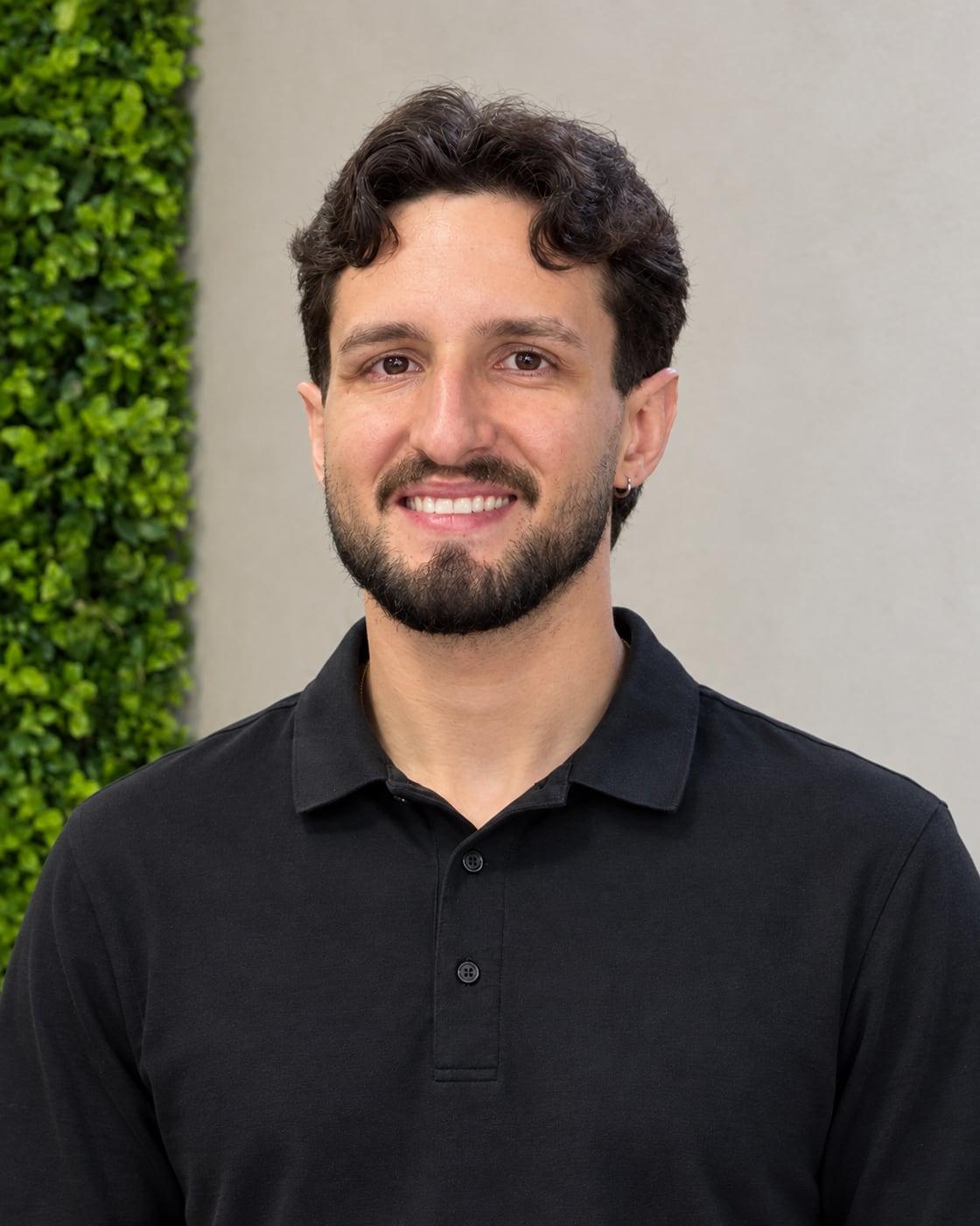 Yuri Melo sorrindo, de polo preta, em frente ao jardim vertical da barbearia