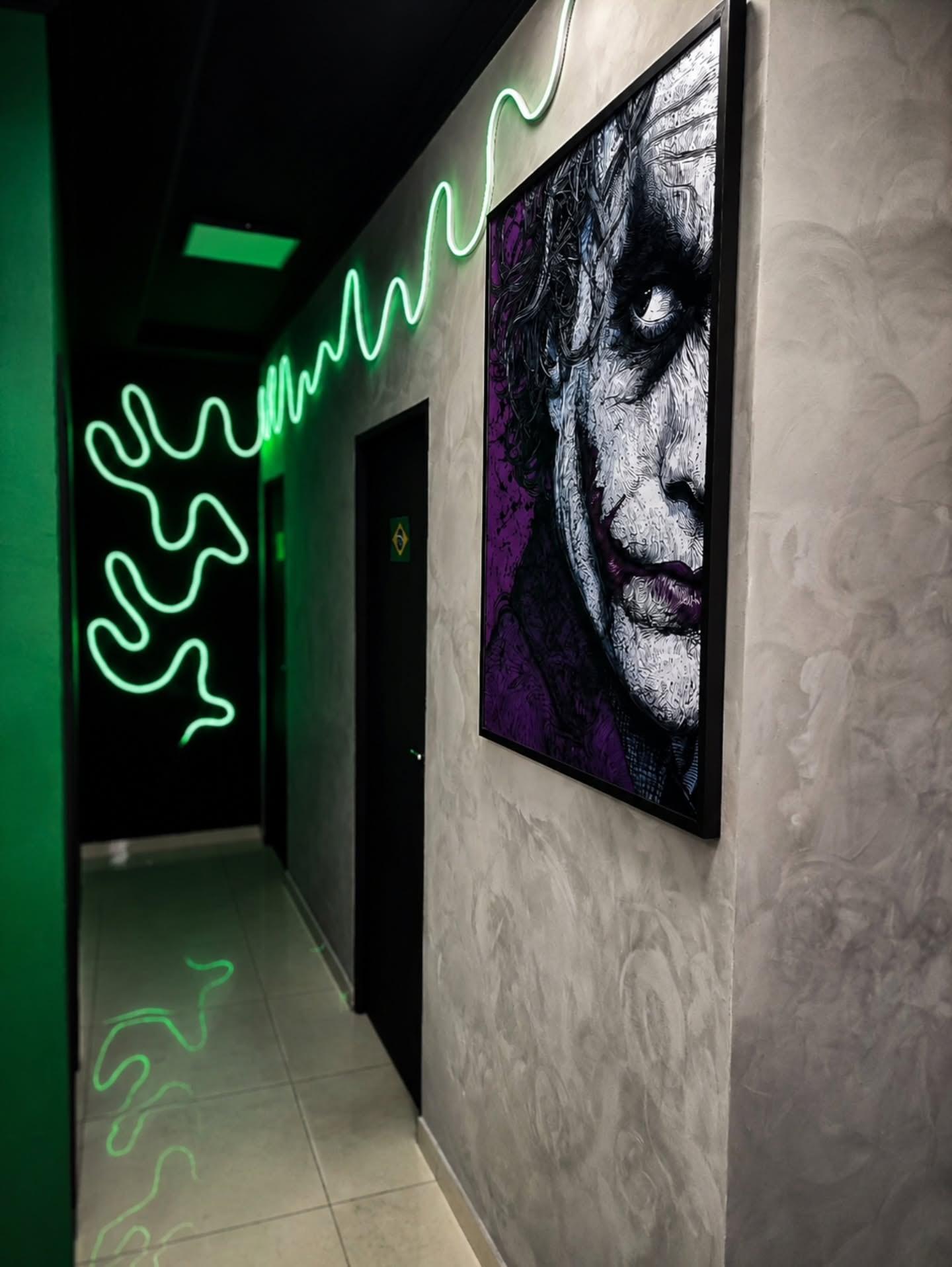 Corredor de cimento iluminado por um neon verde em zigue-zague, com um quadro do Coringa na parede